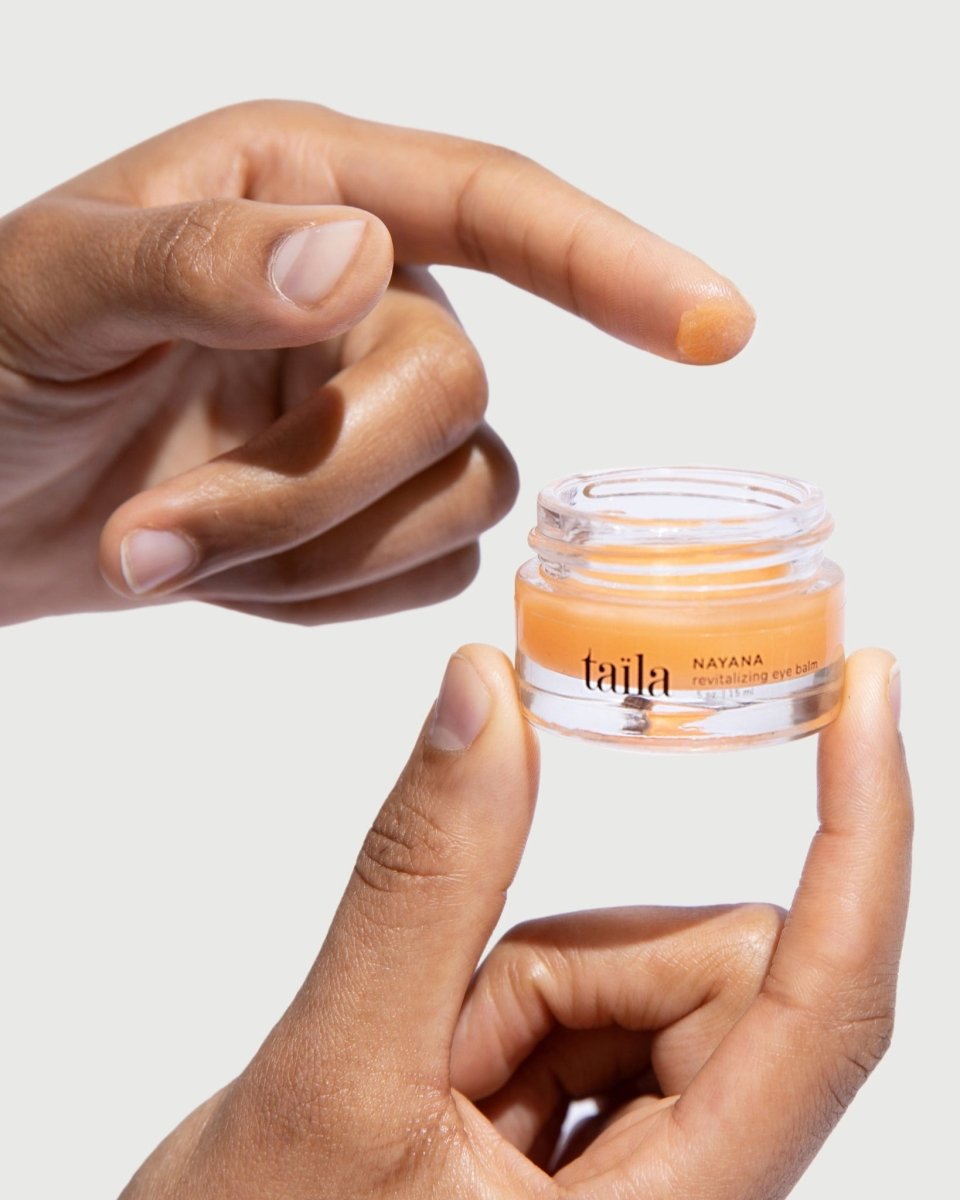 A hand gently holding the taïla NAYANA Revitalizing Eye Balm glass jar against a minimalist white background.