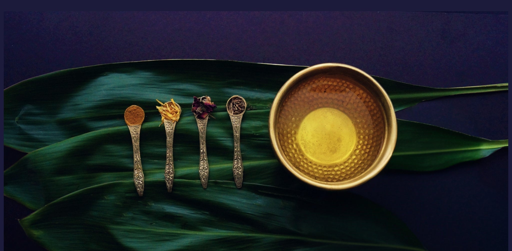 An elevated flat lay of raw, vibrant Ayurvedic botanical ingredients arranged in traditional polished brass spoons alongside a vessel of rich herbal oil, all meticulously presented on a fresh green banana leaf.