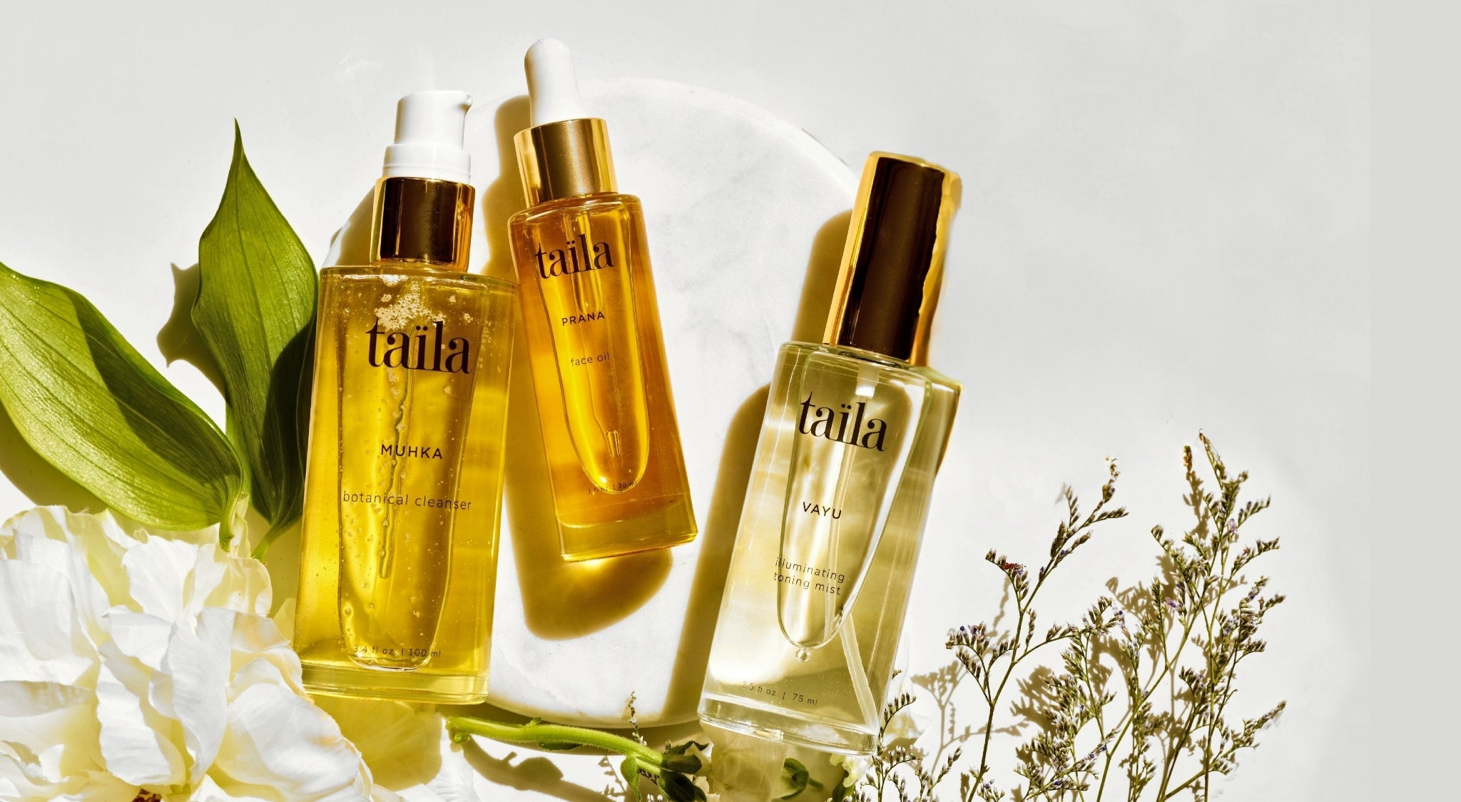 A luxury skincare routine trio featuring a botanical face oil, illuminating toning mist and oil-to-milk cleanser displayed on an elegant marble plate surrounded by fresh blossoms, showcasing a complete taila ritual.