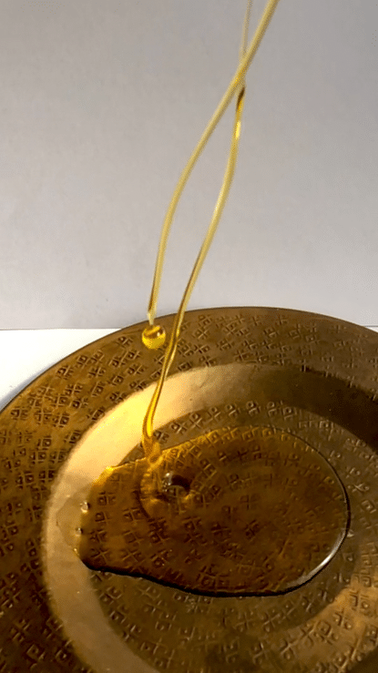 An elegant, looping close-up animation of a single, rich golden drop of Ayurvedic botanical oil slowly pouring from a polished brass vessel plate, capturing the pure, concentrated texture of our active formulas.