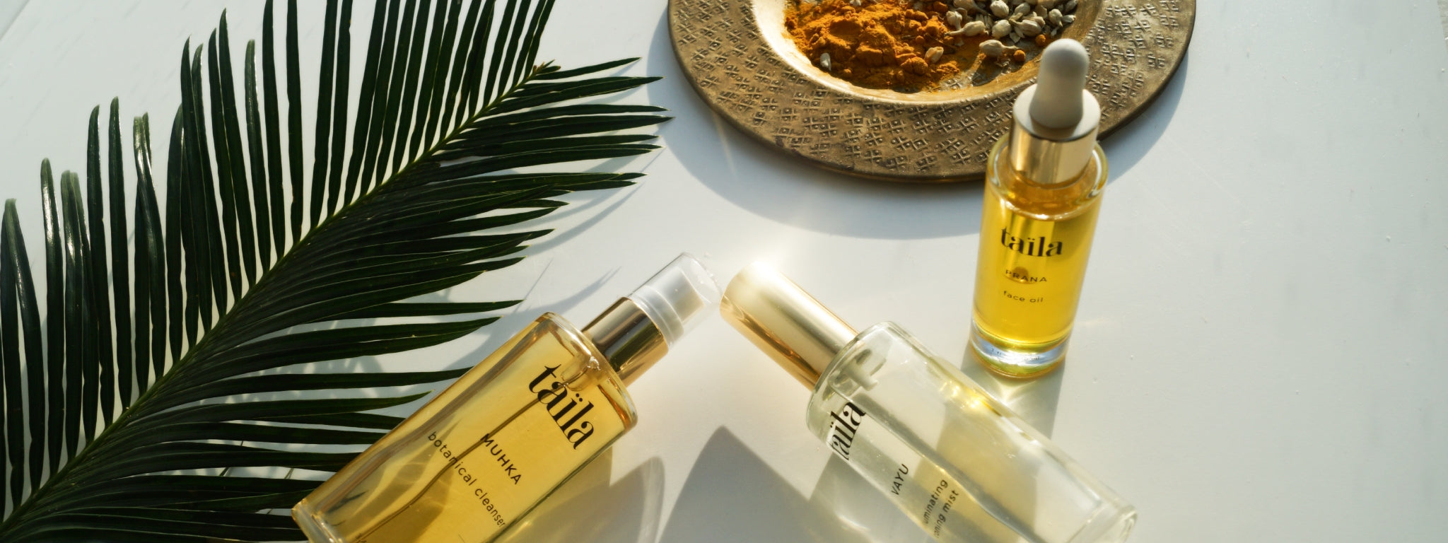 taïla MUHKA cleanser, VAYU mist and PRANA oil bottles displayed with a palm leaf and a brass plate of turmeric and jasmine.