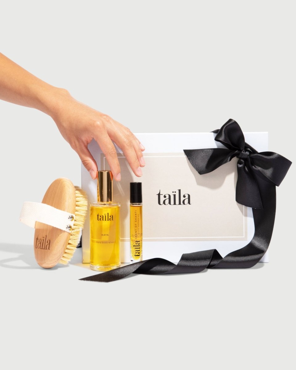 A hand placing taïla KAYA oil, SCENT OF SHANTI aroma oil and a dry brush next to their luxury gift box on a white background.